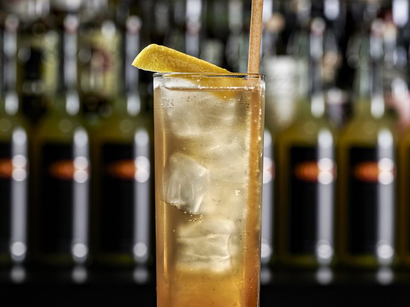 Tall highball glass with ice and an amber cocktail, garnished with a lemon twist and a wooden straw, blurred bar backdrop.