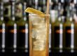 Tall highball glass with ice and an amber cocktail, garnished with a lemon twist and a wooden straw, blurred bar backdrop.