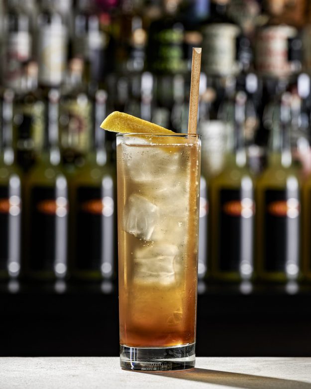 Tall highball glass with ice and an amber cocktail, garnished with a lemon twist and a wooden straw, blurred bar backdrop.