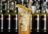 Tall highball glass with ice and an amber cocktail, garnished with a lemon twist and a wooden straw, blurred bar backdrop.