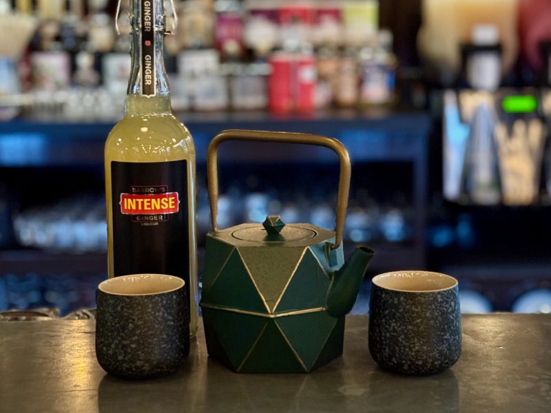 Tea for Two | Barrow's Intense Ginger Liqueur Bottle of Barrows Intense Ginger liqueur on a bar counter, with a green origami centerpiece and two dark cups; blurred bottles in the background.