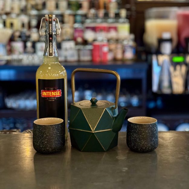 Bottle of Barrows Intense Ginger liqueur on a bar counter, with a green origami centerpiece and two dark cups; blurred bottles in the background.
