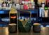 Bottle of Barrows Intense Ginger liqueur on a bar counter, with a green origami centerpiece and two dark cups; blurred bottles in the background.